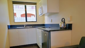 A compact kitchen with white cabinets, a built-in dishwasher, a coffee maker, and a window overlooking a red fence. - Aston Court Motel Blenheim (Blenheim)