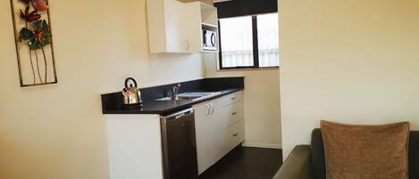 A compact kitchen with a kettle, sink, and white cabinets. There is a window with blinds and a brown cushion on a dark sofa.