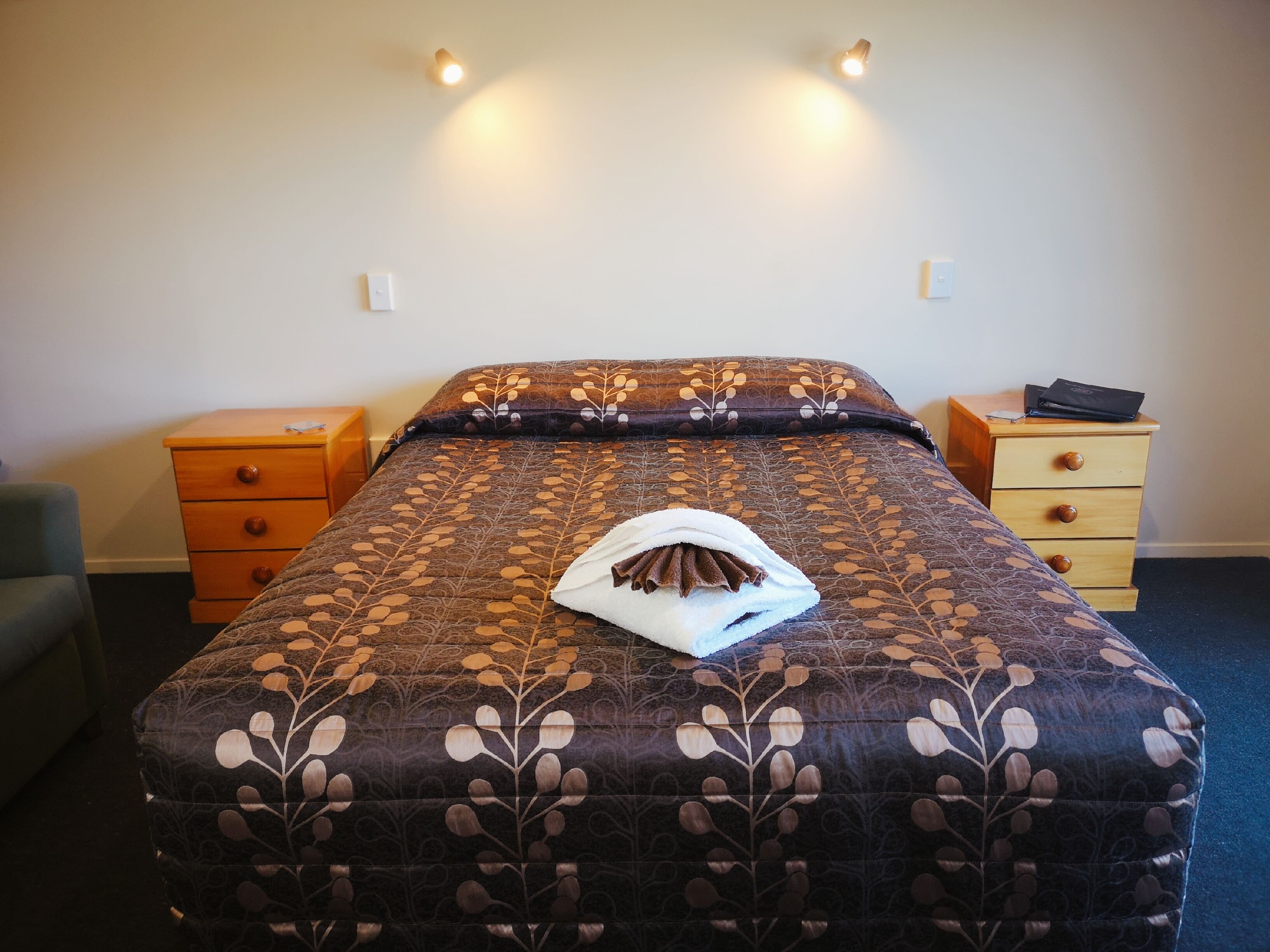 Una cama bien hecha con un edredón estampado, una toalla doblada y un ordenador portátil sobre una mesita de noche de madera.