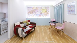 Een gezellige woonkamer met een kleurrijke bank, een klein tafeltje en stoelen. Er is een raam met gordijnen en een aan de muur hangend schilderij.