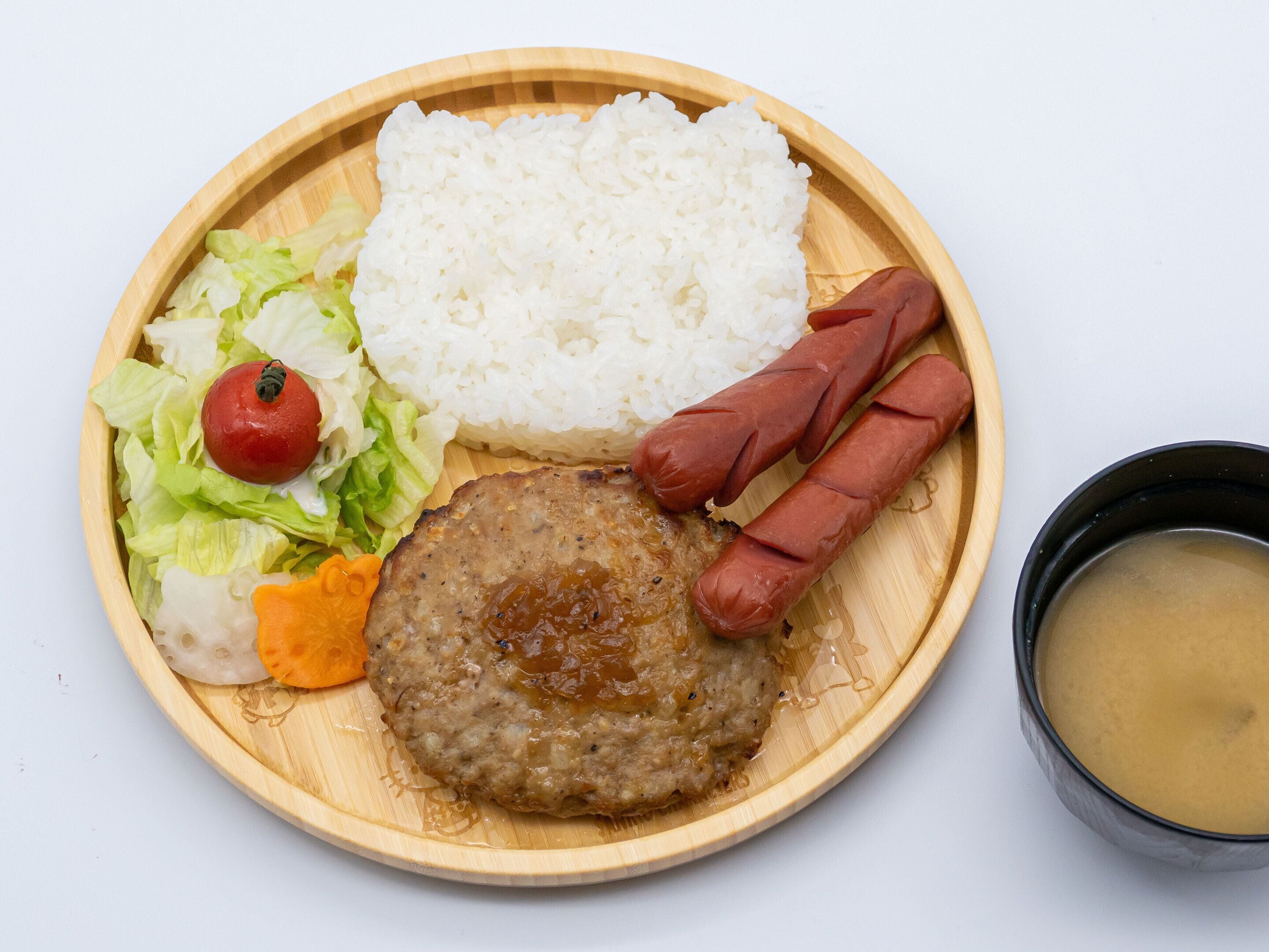 Un plato con arroz blanco, una hamburguesa, salchichas, un tomate, lechuga y un tazón de sopa.