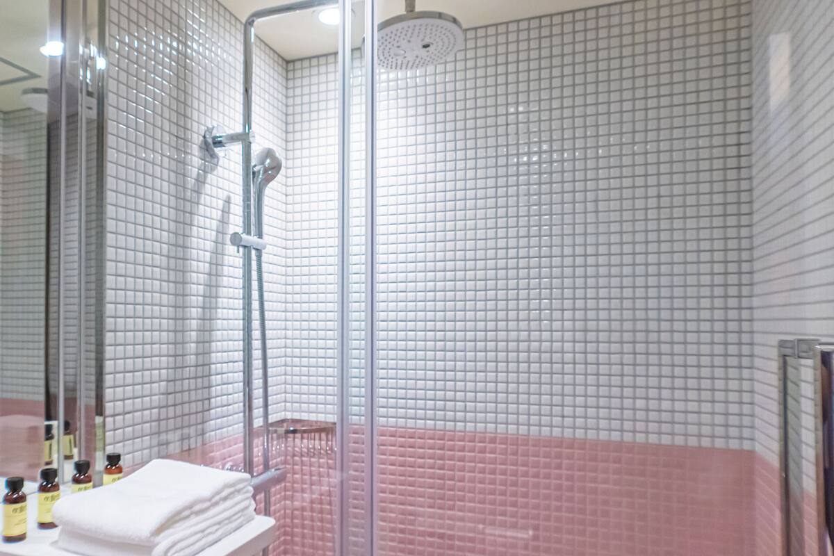 A modern bathroom with a glass shower enclosure, white and pink tiled walls, and a visible bottle of shampoo on the counter.