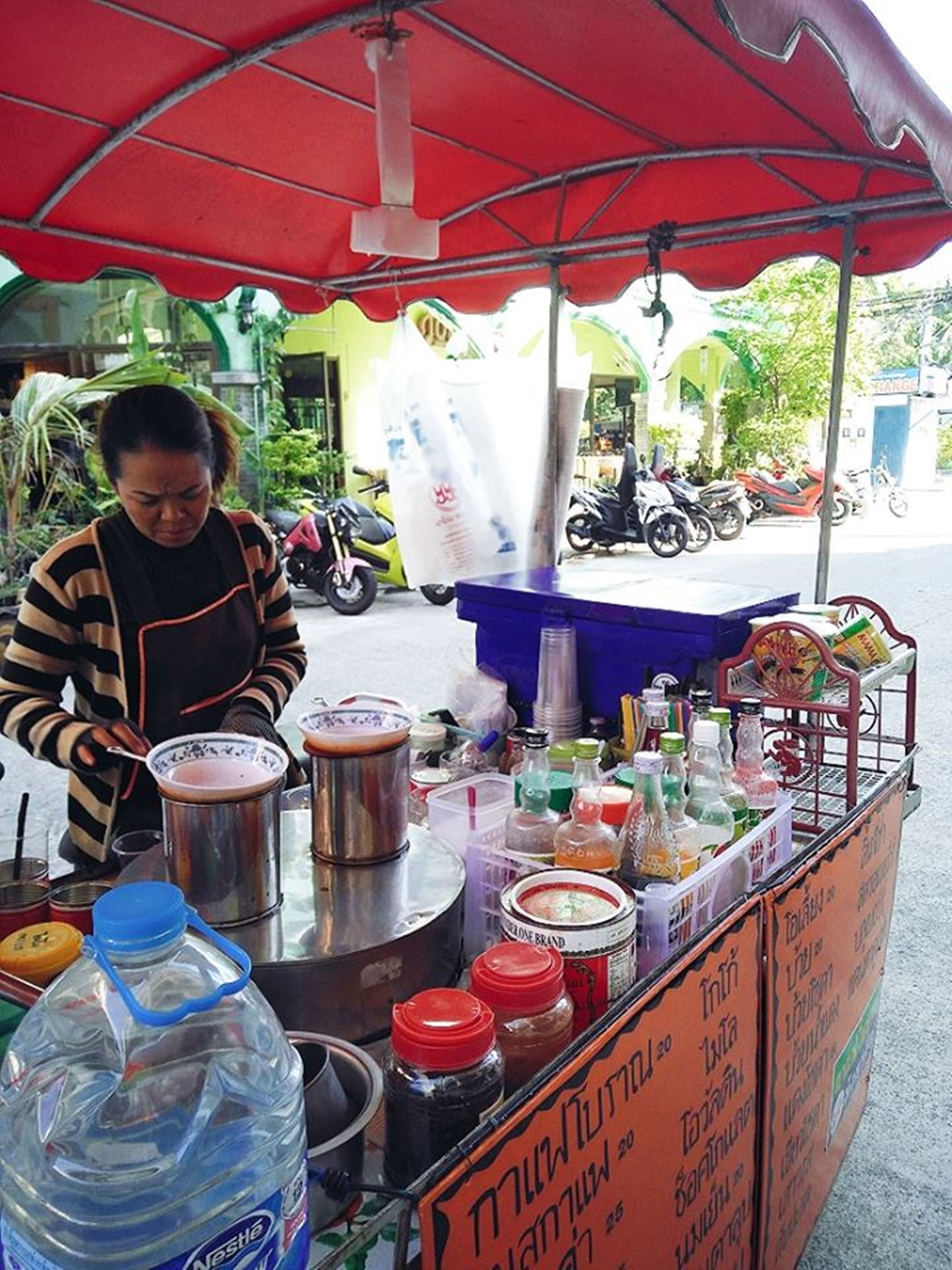 Un étal de vendeur de nourriture de rue proposant une variété de condiments et de boissons, une grande bouteille d’eau et un panneau affichant le menu.