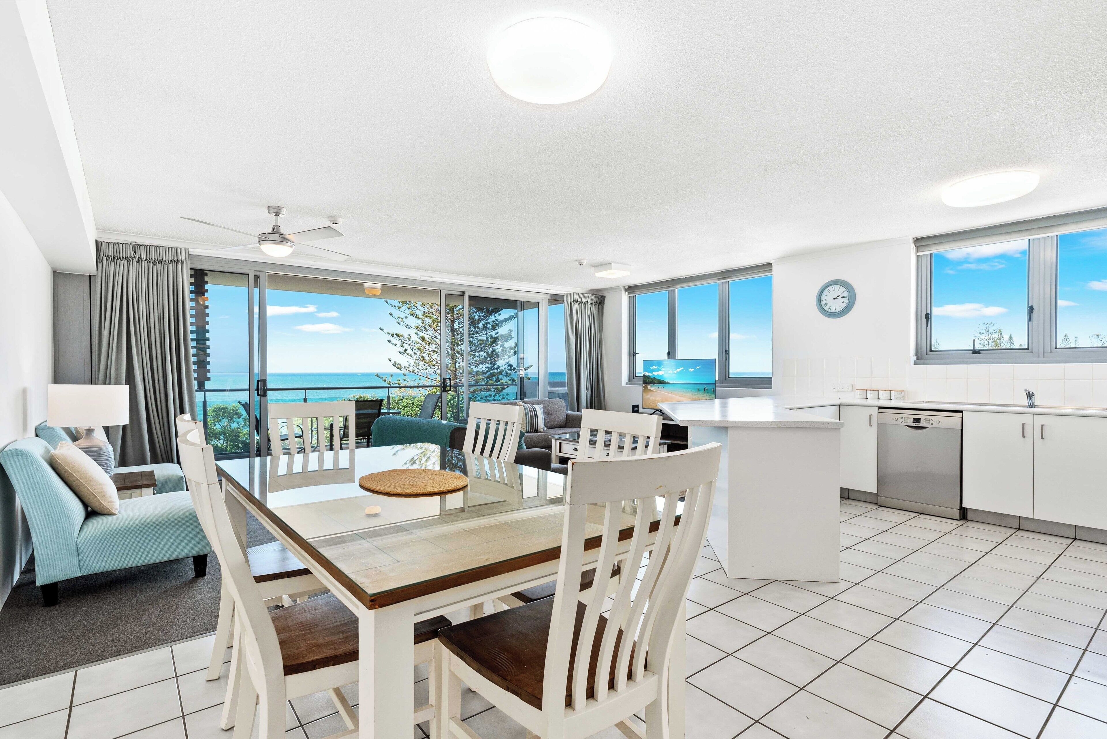 A modern kitchen with a dining area, large windows, and a view of the ocean.