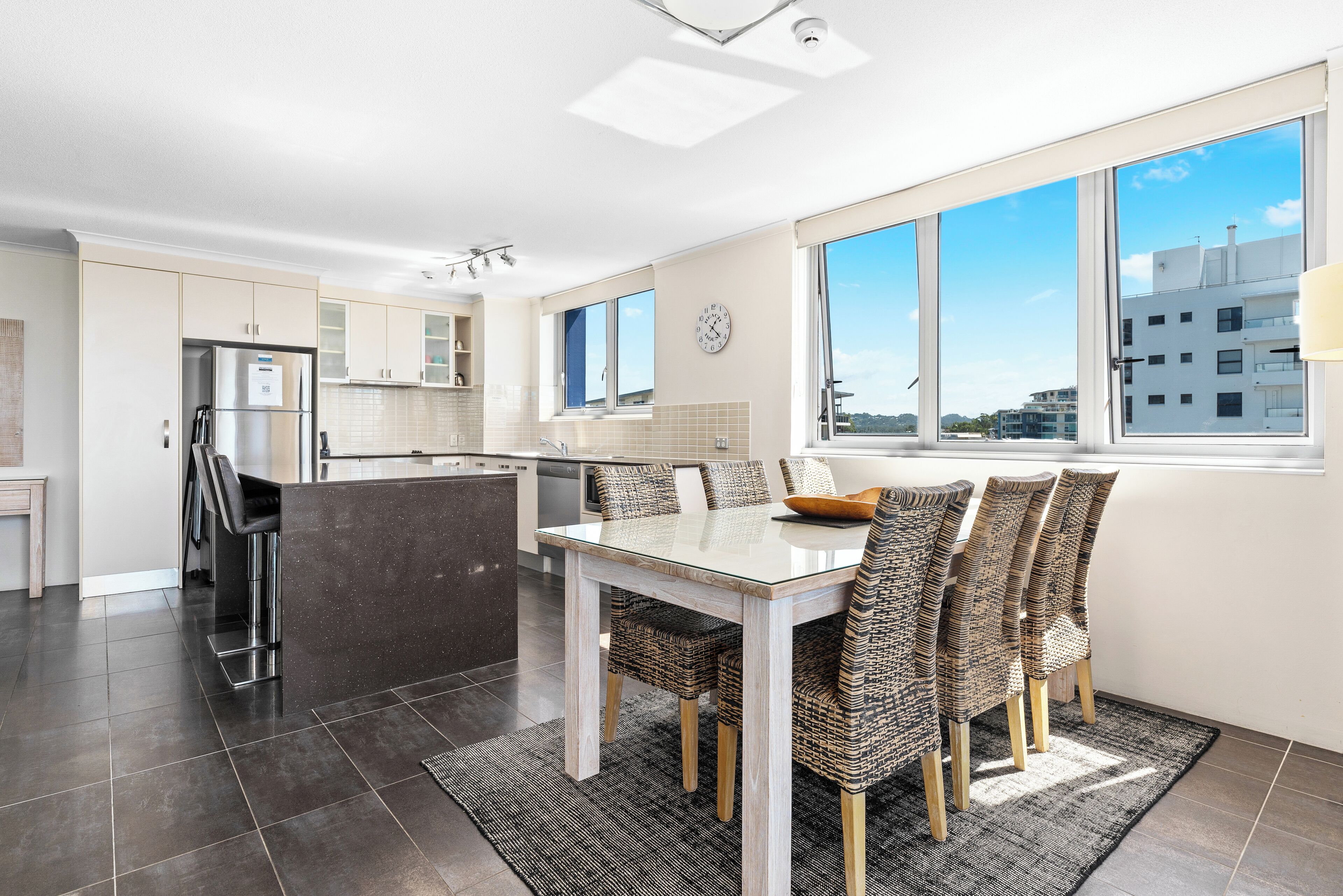 A modern kitchen with a dining area, featuring a large island, dark tiled flooring, and a view of the city through large windows.