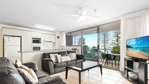 A modern living room with a sofa, coffee table, and a TV. There is a balcony with a view of trees and a beach. - Northwind Holiday Apartments Mooloolaba (Mooloolaba)