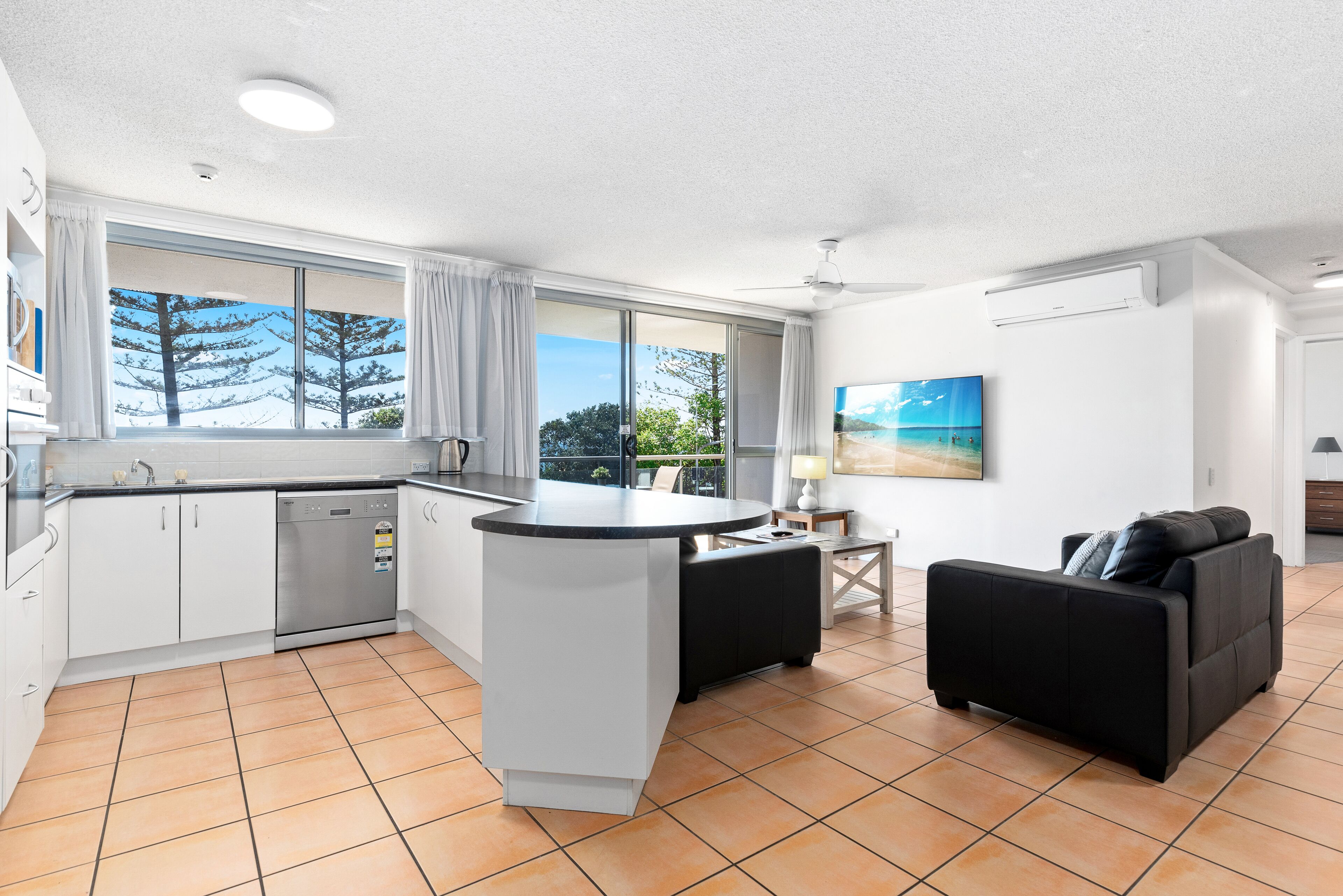 A modern kitchen with a central island, a dining area with a black sofa, and large windows offering a view of trees and sky.