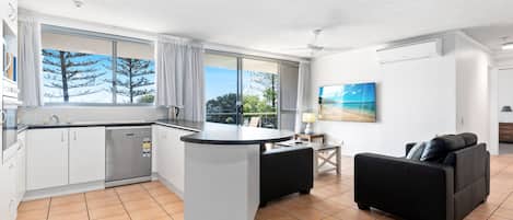 A modern kitchen with a central island, a dining area with a black sofa, and large windows offering a view of trees and sky.