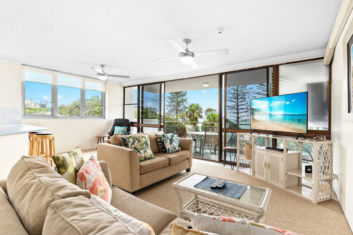 A spacious living room with a large window, a ceiling fan, a flat-screen TV, and a comfortable seating area.