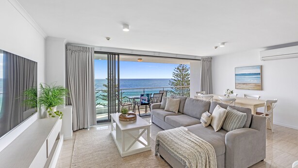 A modern living room with a large sofa, a coffee table, and a dining area. - Northwind Holiday Apartments Mooloolaba (Mooloolaba)
