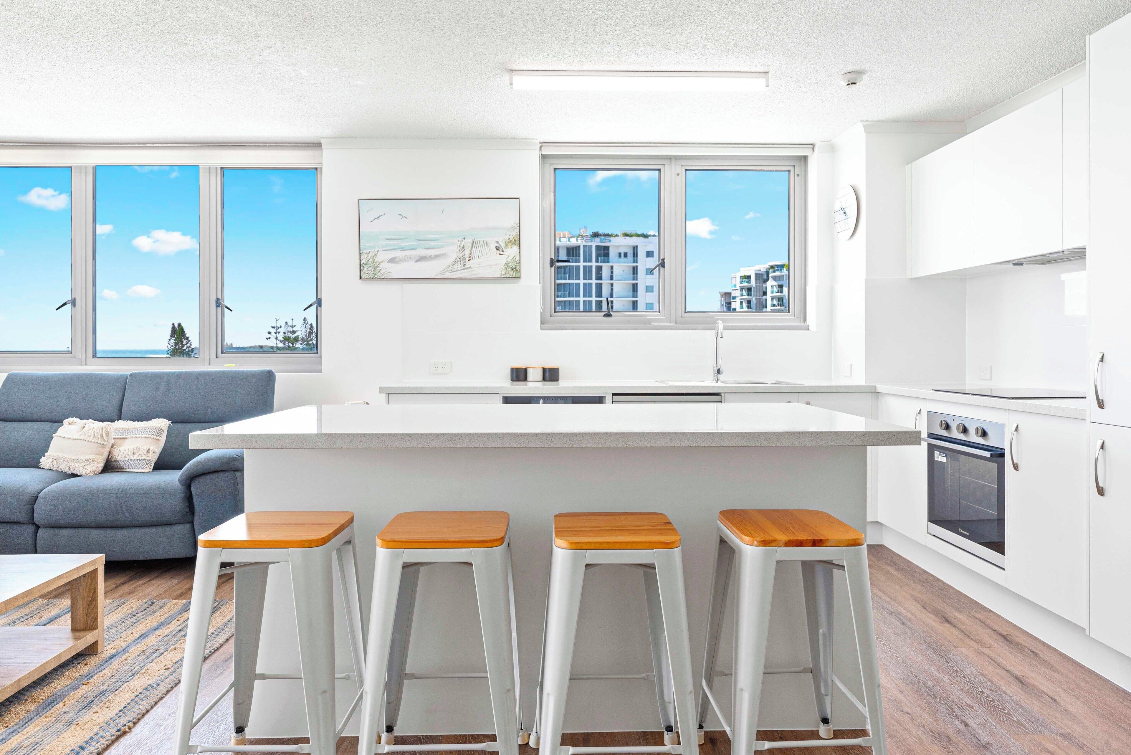 A modern kitchen with white cabinets, a central island with stools, and large windows offering a view of buildings and trees.
