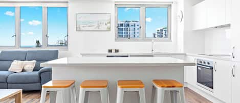 A modern kitchen with white cabinets, a central island with stools, and large windows offering a view of buildings and trees.