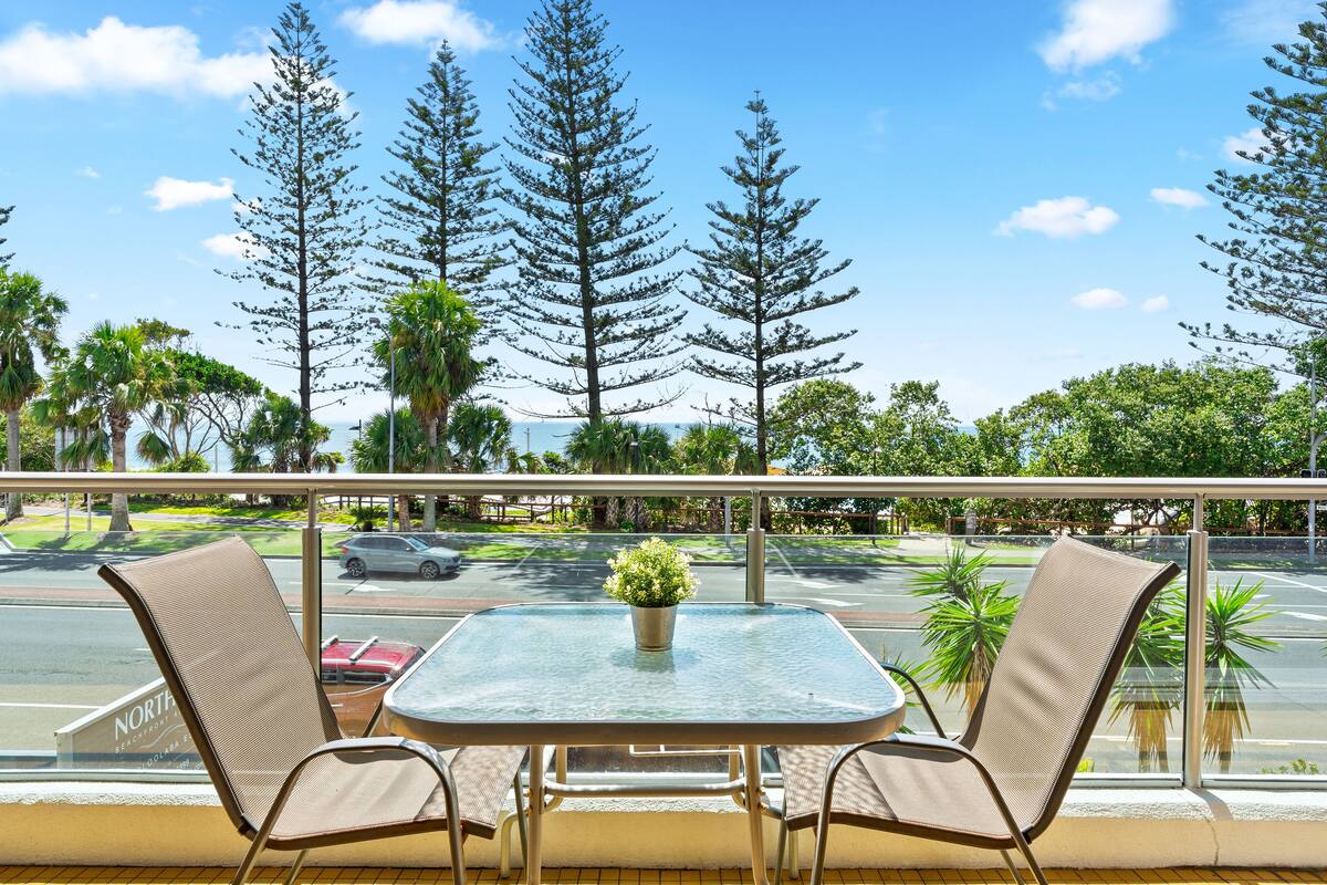 A balcony with a table and chairs, overlooking a street with cars and tall trees.