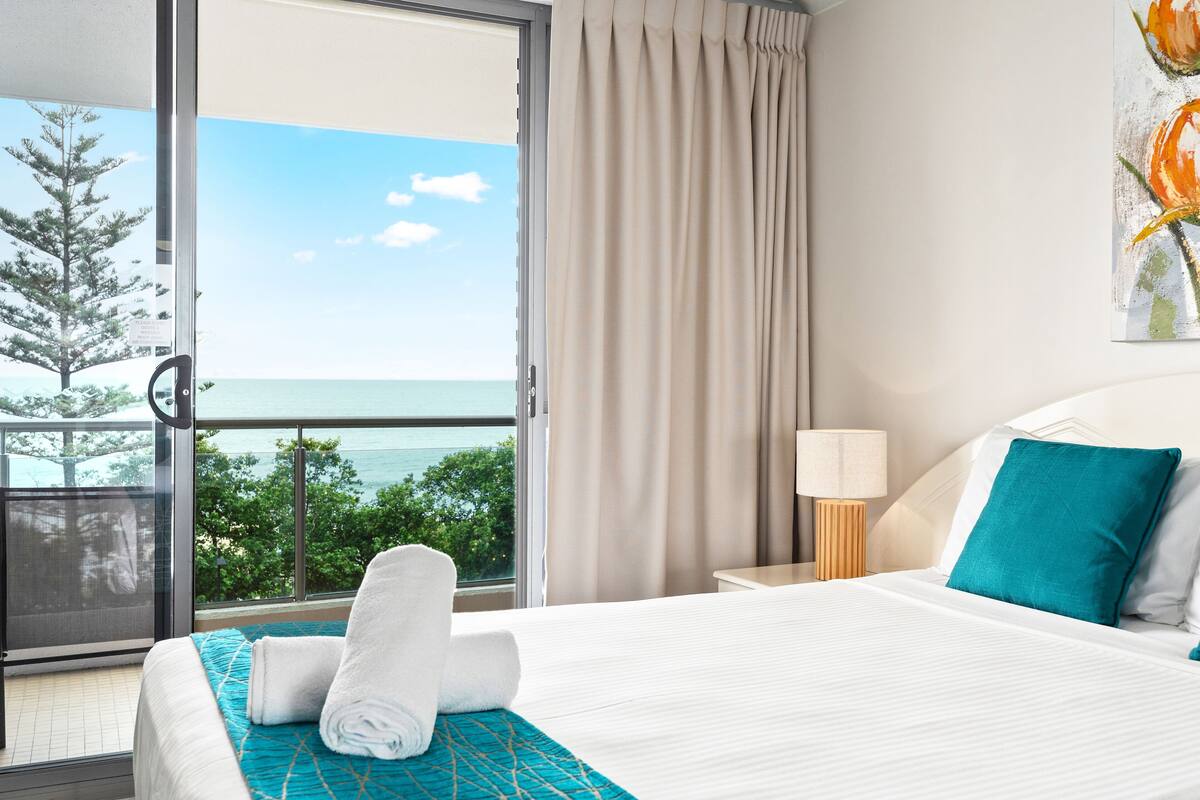 A hotel room with a bed, a nightstand, a lamp, and a view of the ocean through a large window.