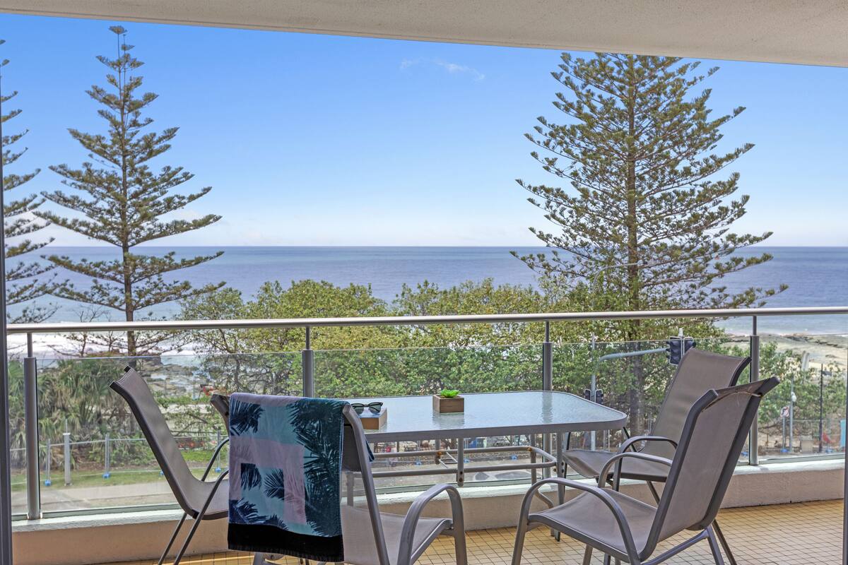 A balcony with a table and chairs, overlooking a beach and ocean.