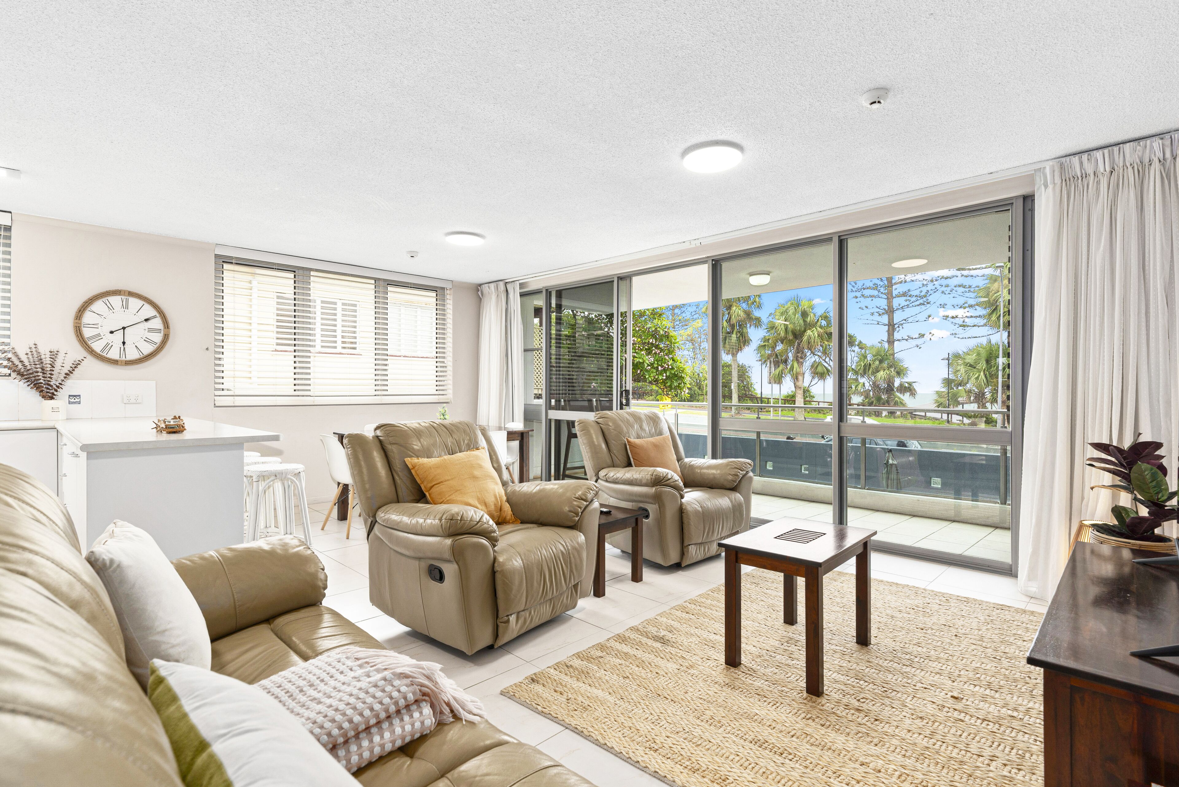 Ein modernes Wohnzimmer mit Ledersofas, einem Holztisch und einer großen Glasschiebetür, die auf einen Balkon mit Blick auf Palmen führt.