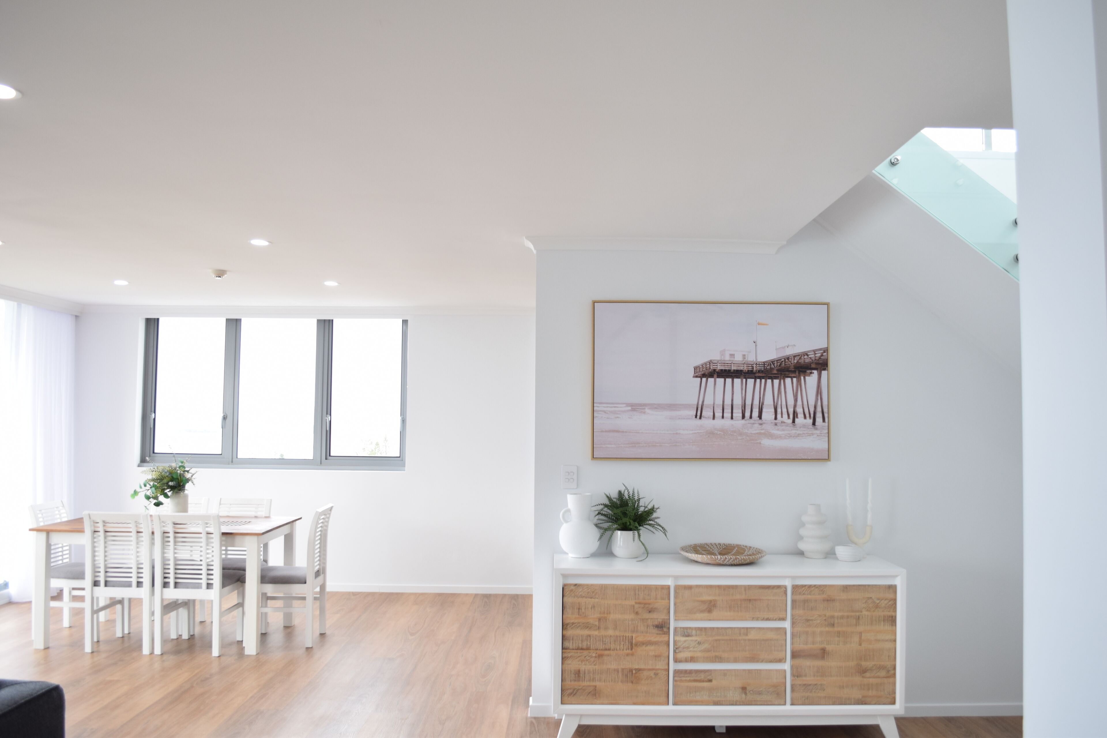 Ein moderner Essbereich mit einem weißen Tisch und Stühlen, einem Holzschrank mit Dekoration und einem großen Fenster, das für natürliches Licht sorgt.