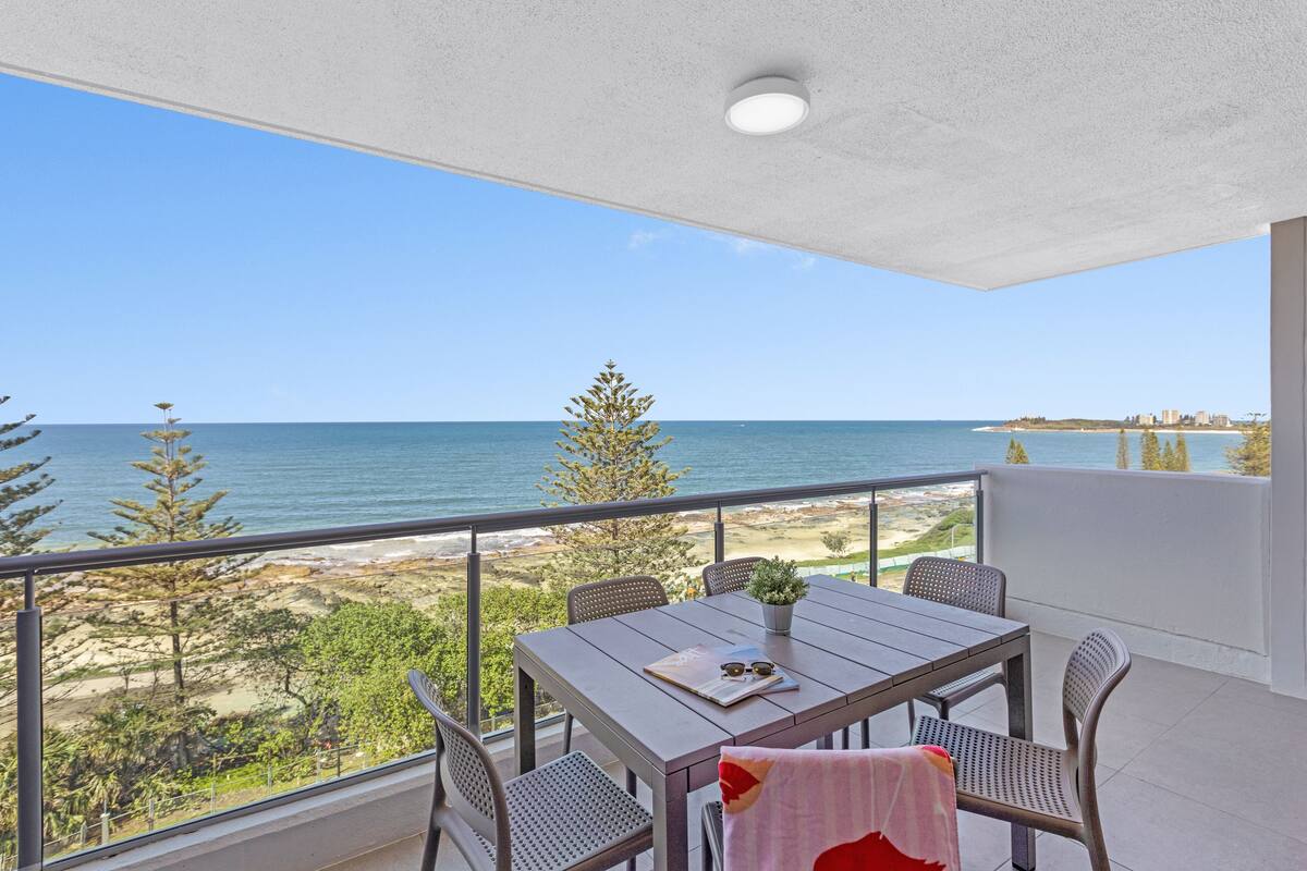 A balcony with a table, chairs, and a potted plant, overlooking a beach and ocean.