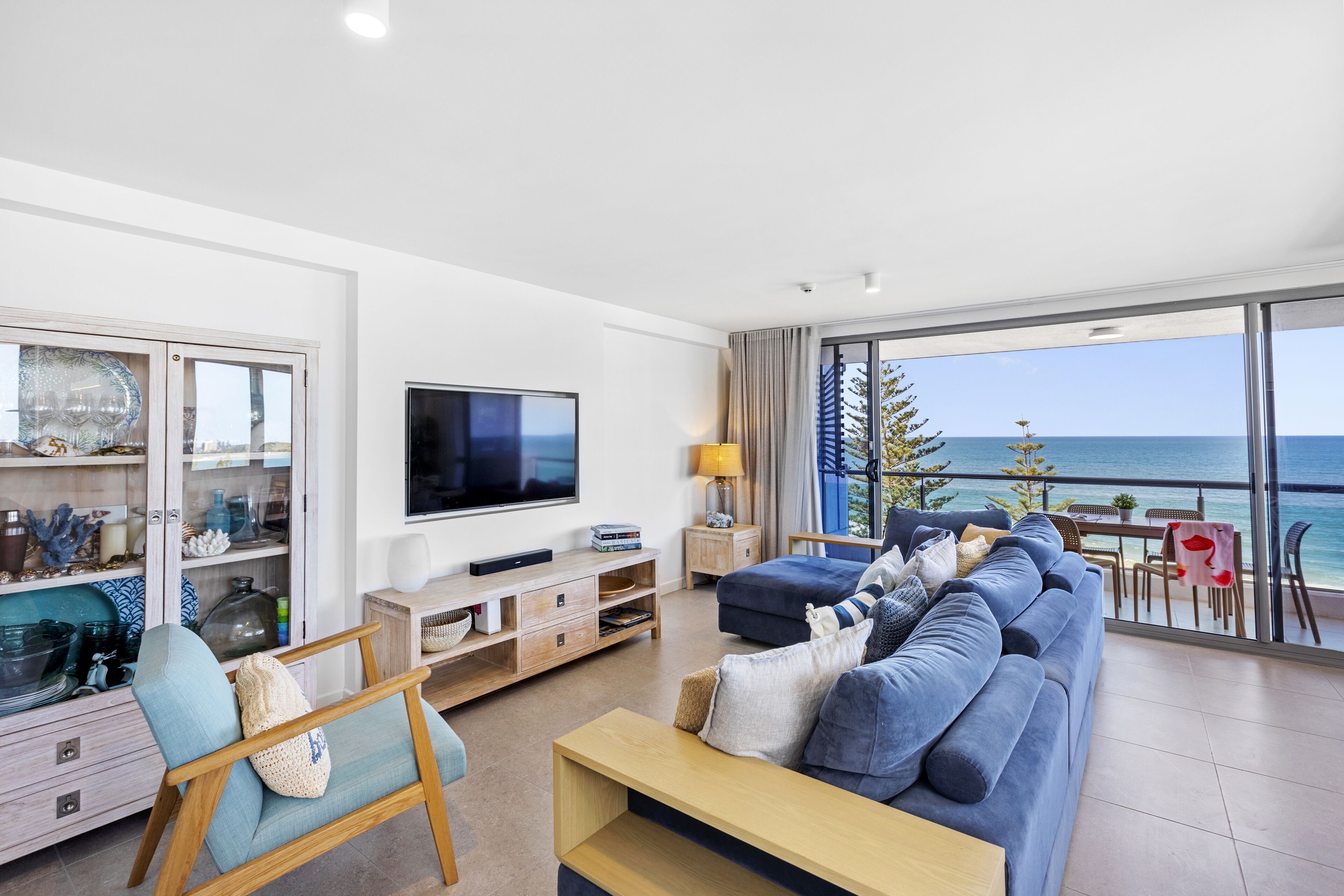 A modern living room with a large TV, comfortable seating, a glass cabinet with decorative items, and a view of the ocean.