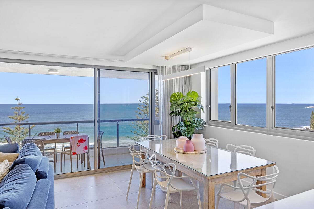 A modern living room with a glass dining table, white chairs, a blue sofa, and a view of the ocean.
