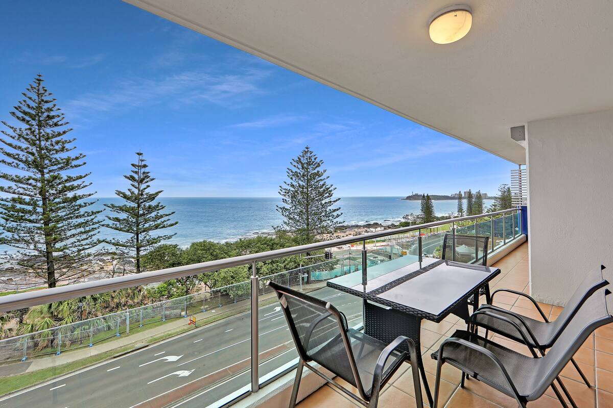 A balcony with a table and chairs overlooking a coastal road and ocean.