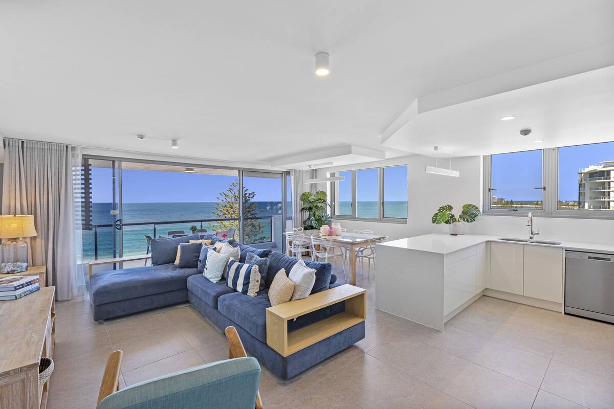 A modern living room with a large sofa, a dining area, and a kitchen with a view of the ocean.