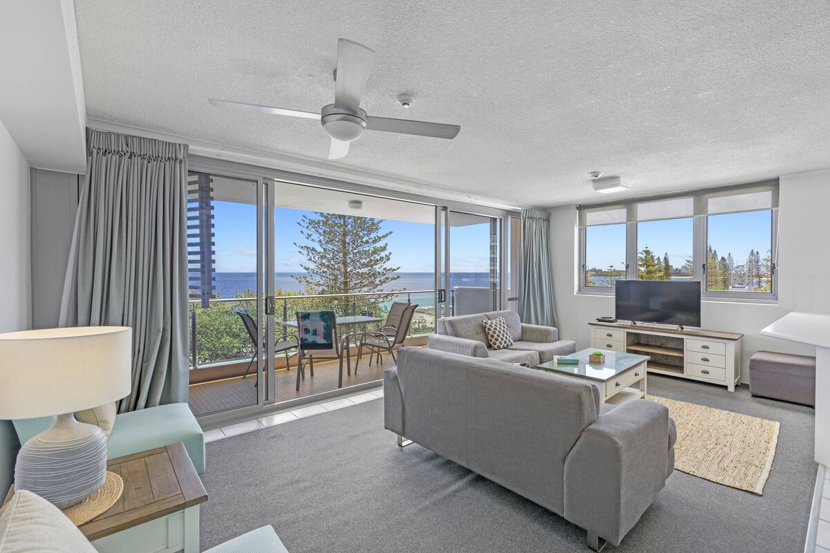 A modern living room with a grey sofa, a TV, and a balcony with a view of the sea.