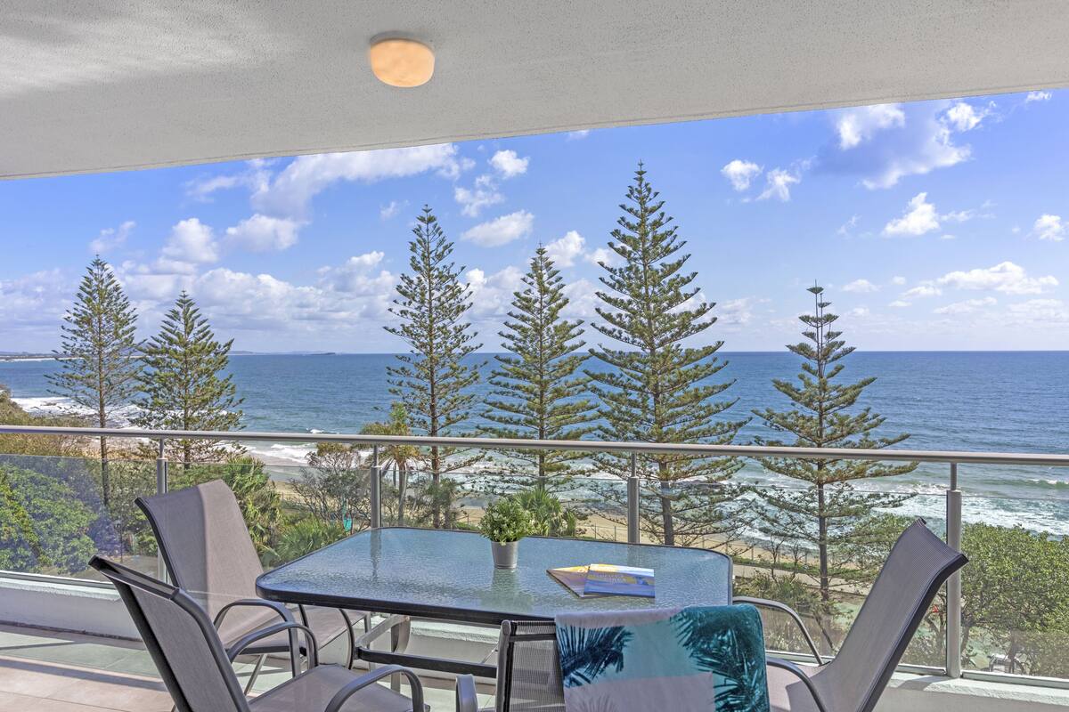 A balcony with a glass table, two chairs, and a view of the ocean and trees.