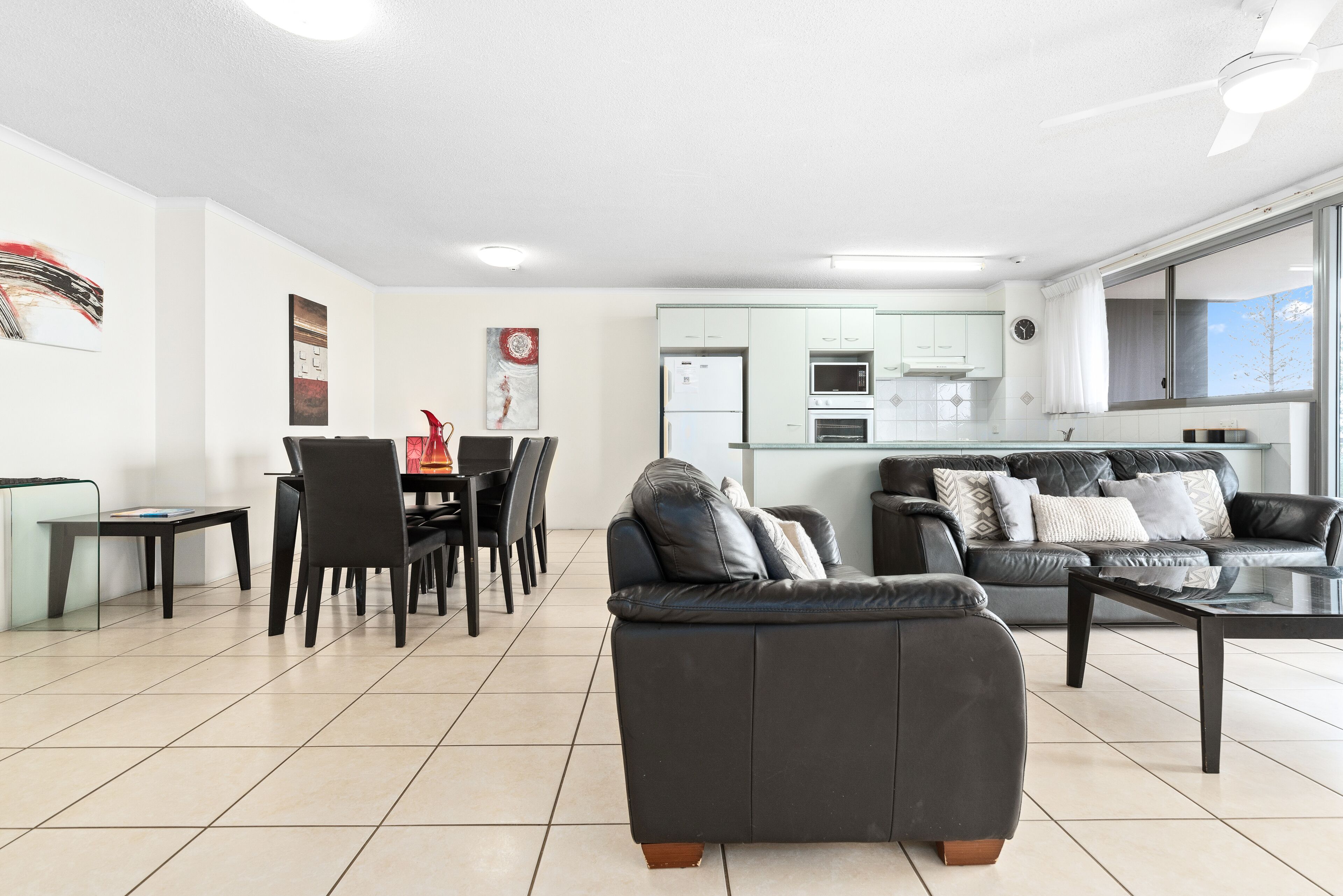 A modern living room with a black leather sofa, a dining area with a table and chairs, and a kitchen with white cabinets and appliances.