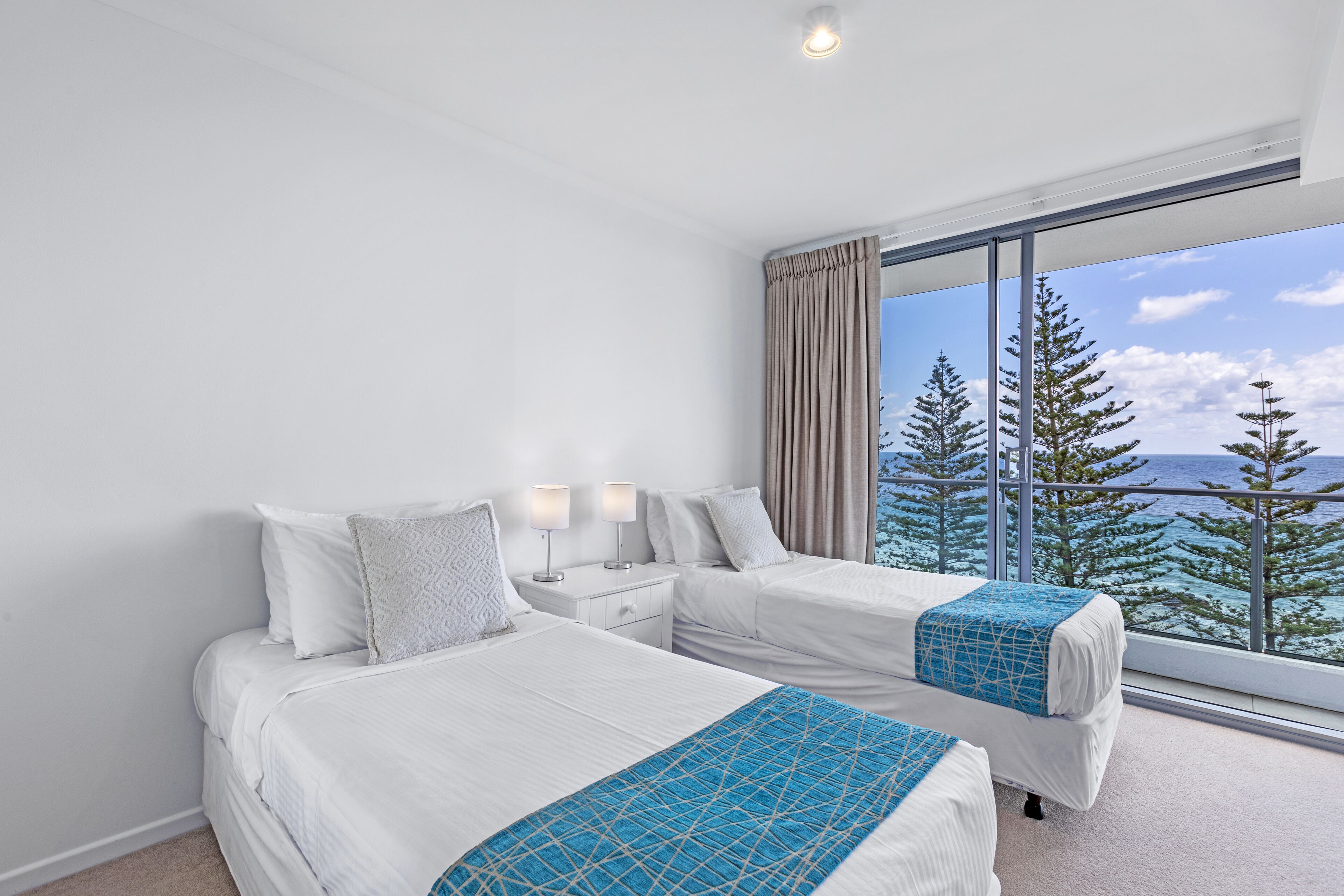 Habitación de hotel con dos camas, un ventanal con vistas a árboles y al mar, y un balcón.
