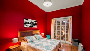 A bedroom with a red wall, a bed with a quilt, a nightstand, a chair, and a window with curtains. - La Chimba Hostel (Santiago)