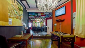 A room with a wooden table, chairs, a chandelier, a foosball table, and a television on the wall. - La Chimba Hostel (Santiago)
