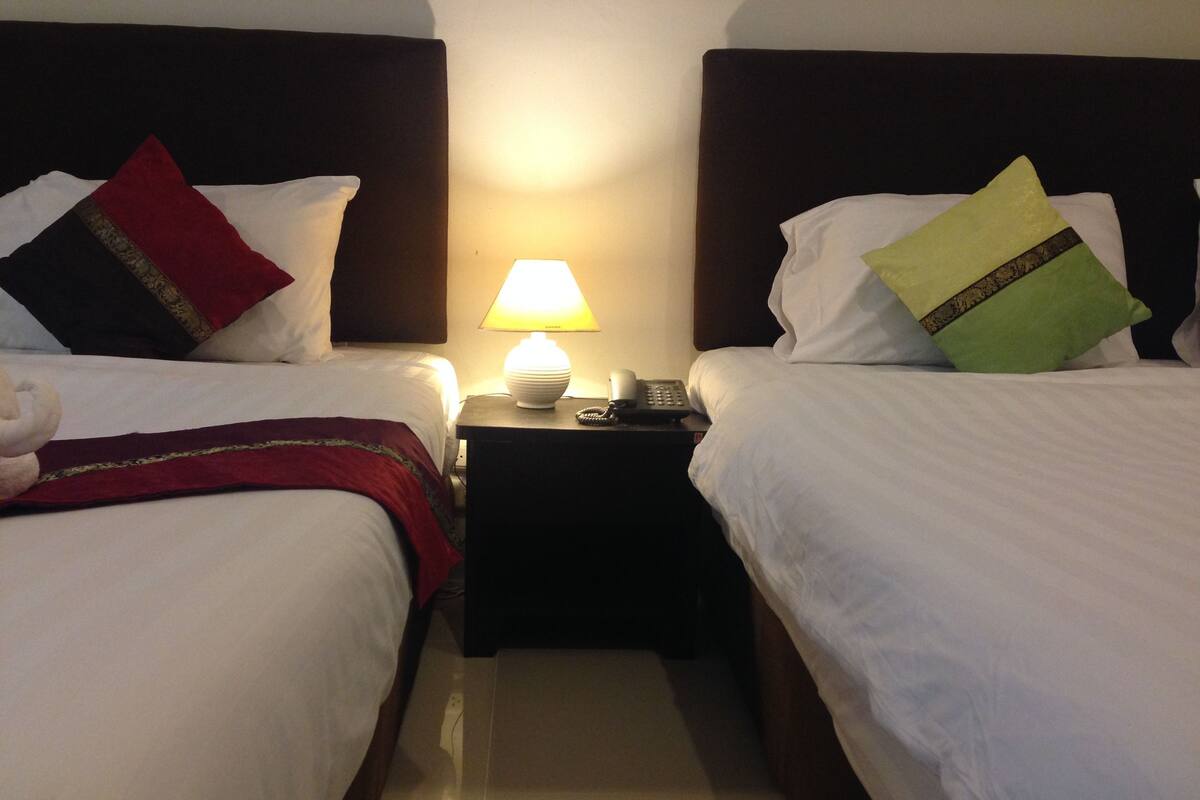 Een hotelkamer met twee bedden, elk voorzien van een kussen en dekbed. Er is een nachtkastje met een lamp en telefoon.