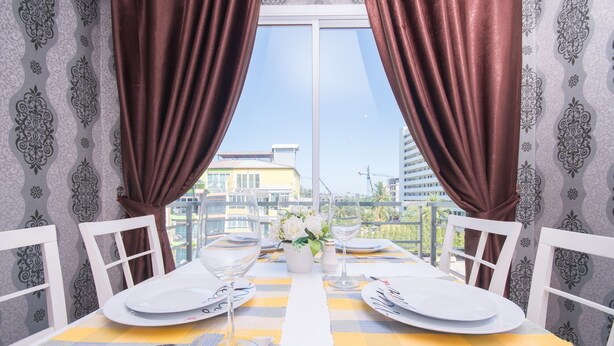 A dining table set with white plates, cutlery, and a glass of water, with a view of buildings through a window with brown curtains. - Grande Elegance Serviced Apartment (Patong)