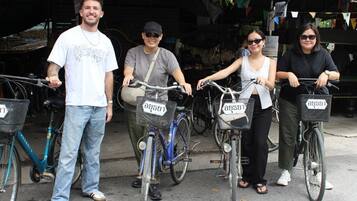 Four people standing beside bicycles with baskets, in an outdoor setting with a covered area and trees in the background.