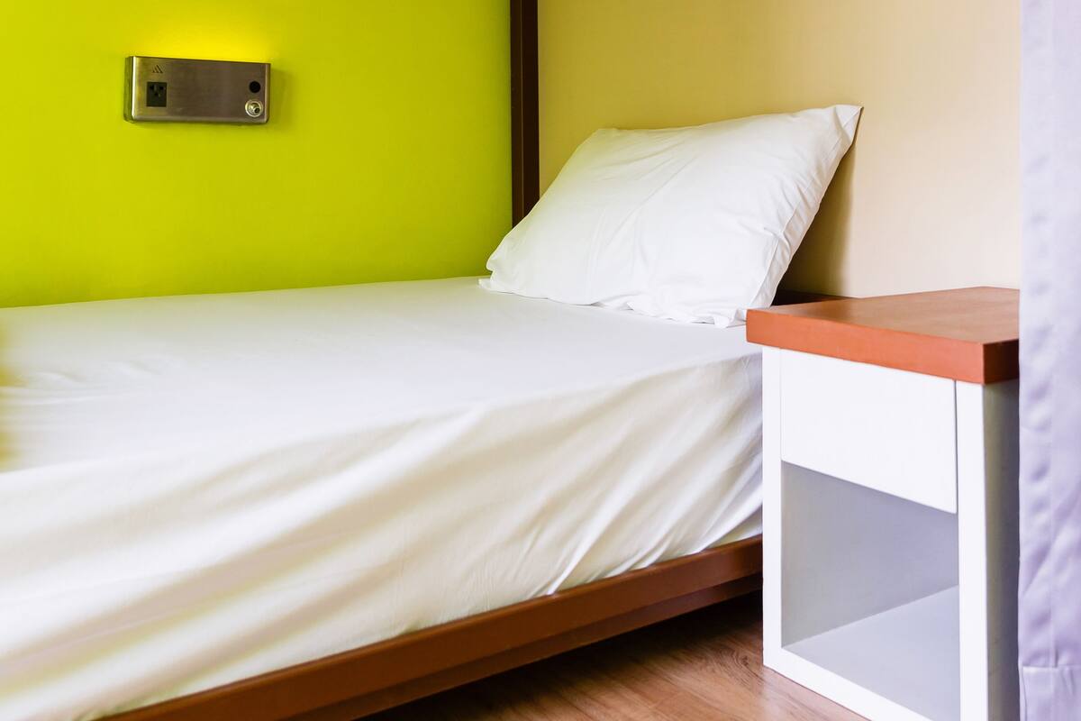 A single bed with a wooden frame, a white mattress, and a white pillow. There is a small bedside table with a lamp and a wall-mounted light switch. The room has a yellow wall and a wooden ceiling with exposed beams.