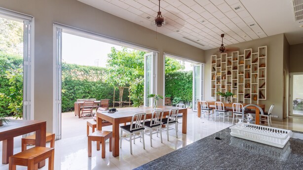 A spacious dining area with a large table and chairs, a kitchen island, and a shelving unit filled with dishes. - Siamaze Hostel (Bangkok)