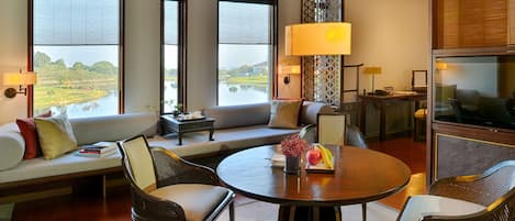 A spacious living room with a sofa, chairs, a round table, and a TV. Large windows offer a view of a lake and greenery.
