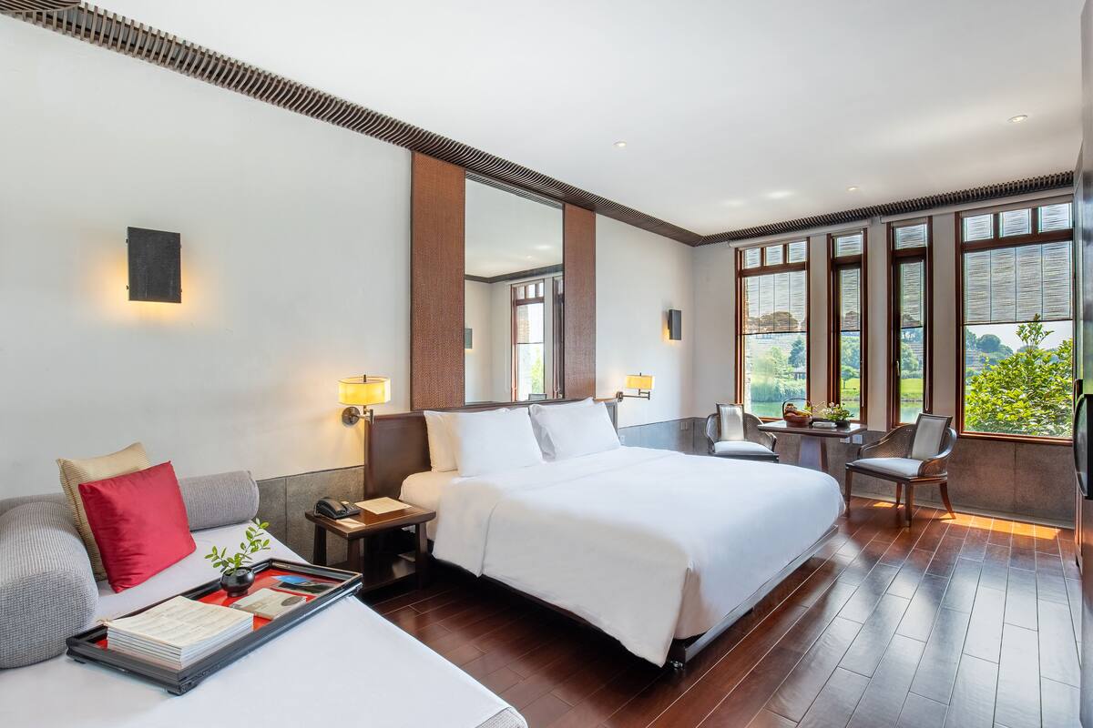 Quarto de hotel moderno com uma cama grande, um sofá, uma mesinha de centro com uma luminária e vista para o verde através de amplas janelas.