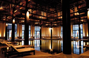 Une vaste piscine intérieure, dotée de chaises longues et de piliers, et entourée de grandes fenêtres.