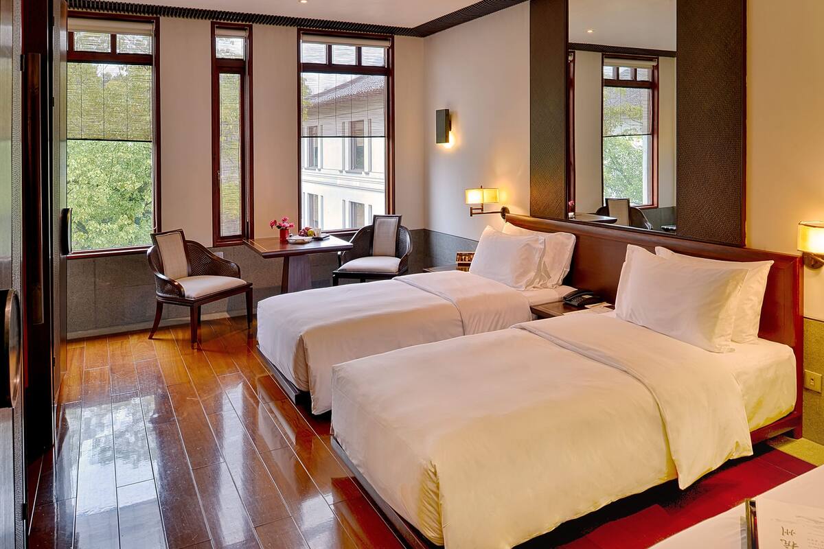 Quarto de hotel com duas camas, um espaço de estar com uma poltrona e mesa, e janelas amplas com vista para o exterior.
