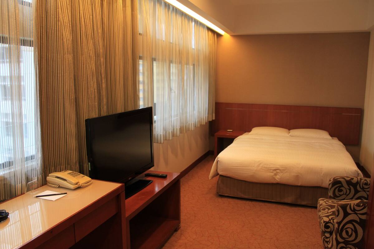 A hotel room with a bed, a desk with a television, a telephone, and a chair.