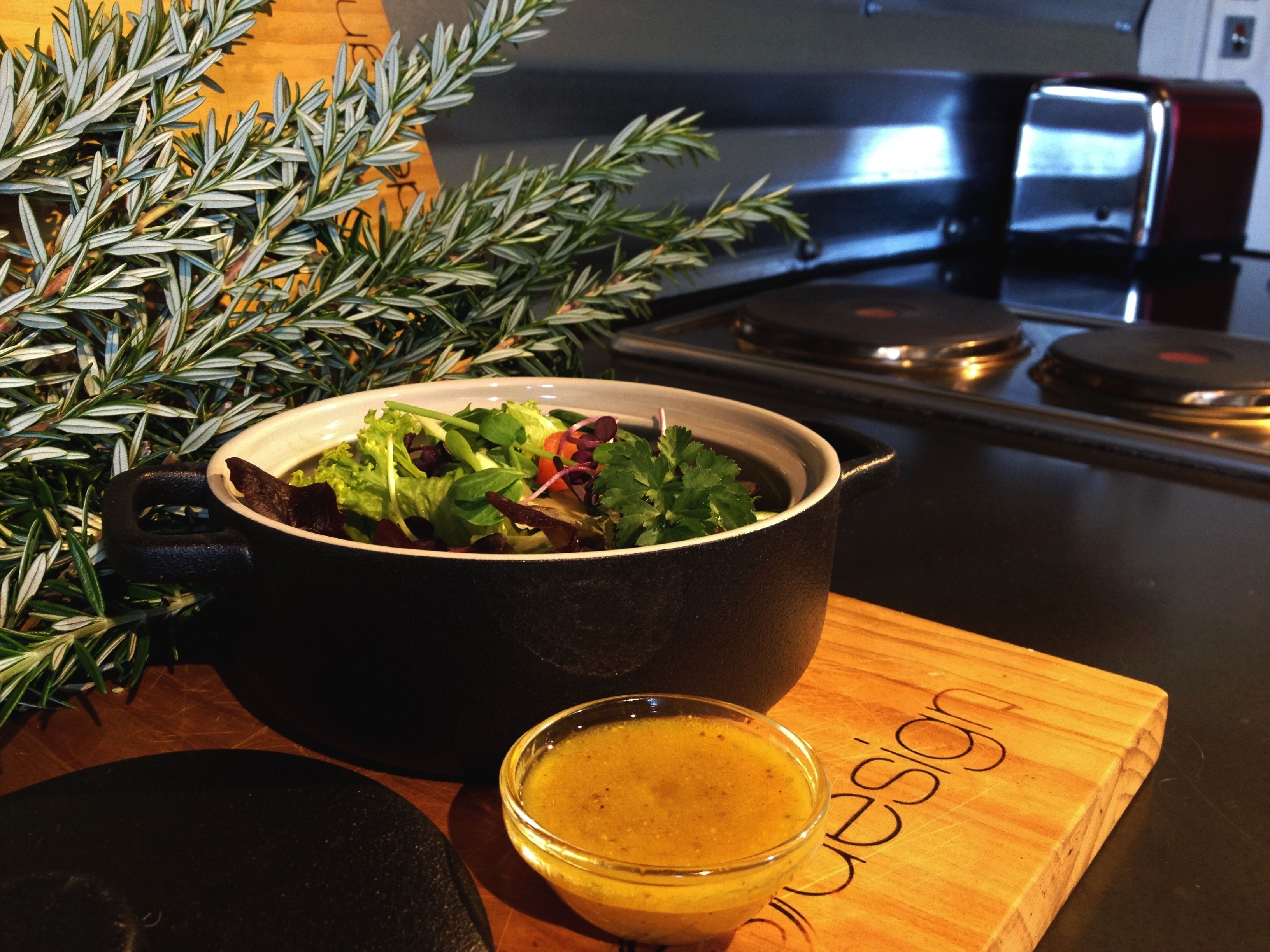 A bowl of salad on a wooden board, a glass of sauce, and a plant with green leaves.