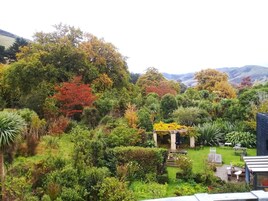 A lush garden with a variety of trees and shrubs, a small patio area with outdoor furniture, and a view of distant hills.