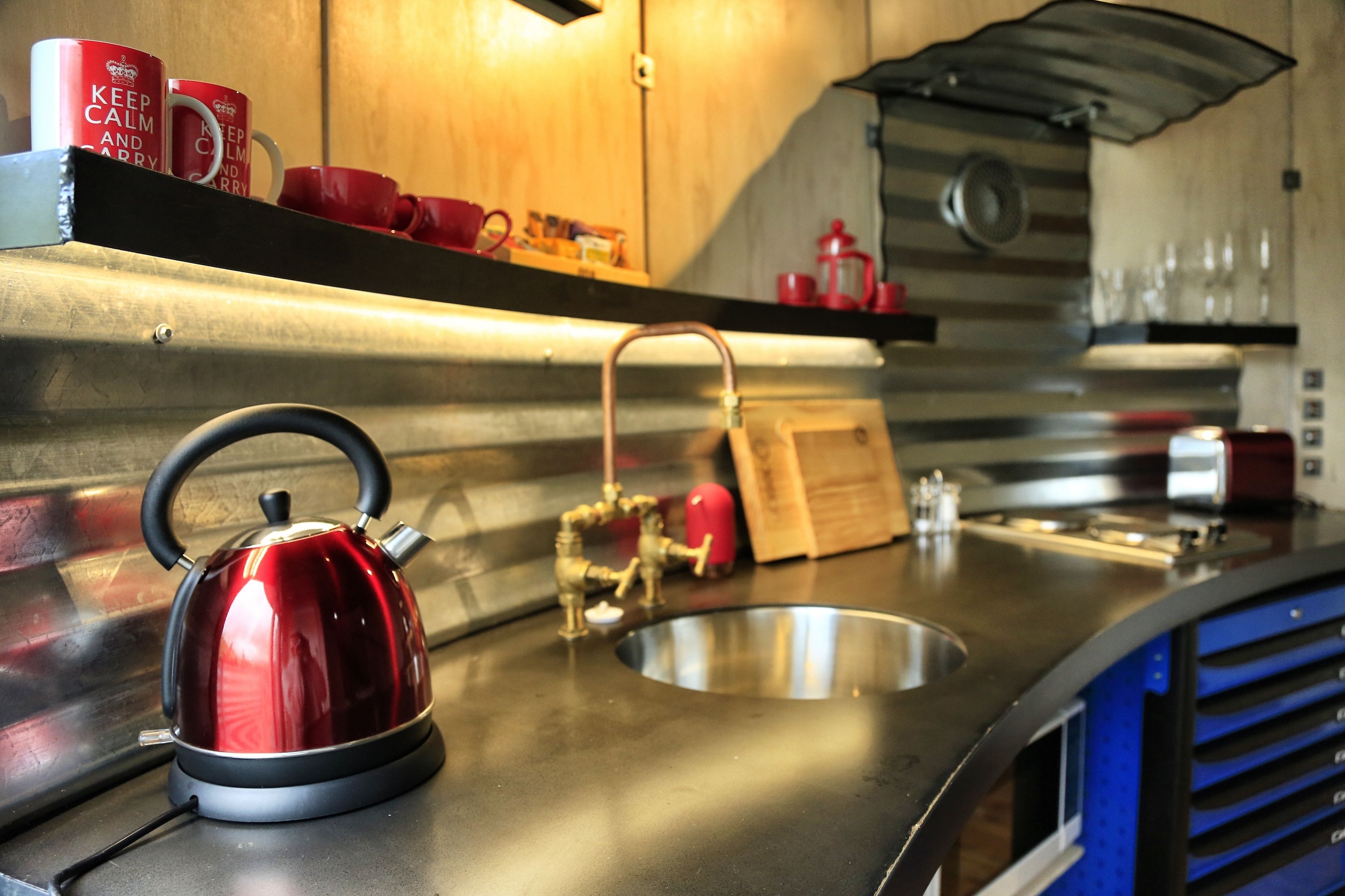 A stainless steel kitchen with a kettle, a sink, and red mugs on a shelf.