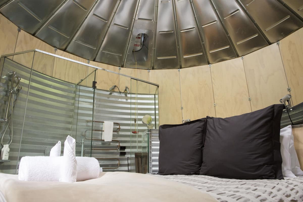A modern hotel room with a curved ceiling, a glass shower enclosure, a bed with dark and white pillows, and a neatly made bed with towels.