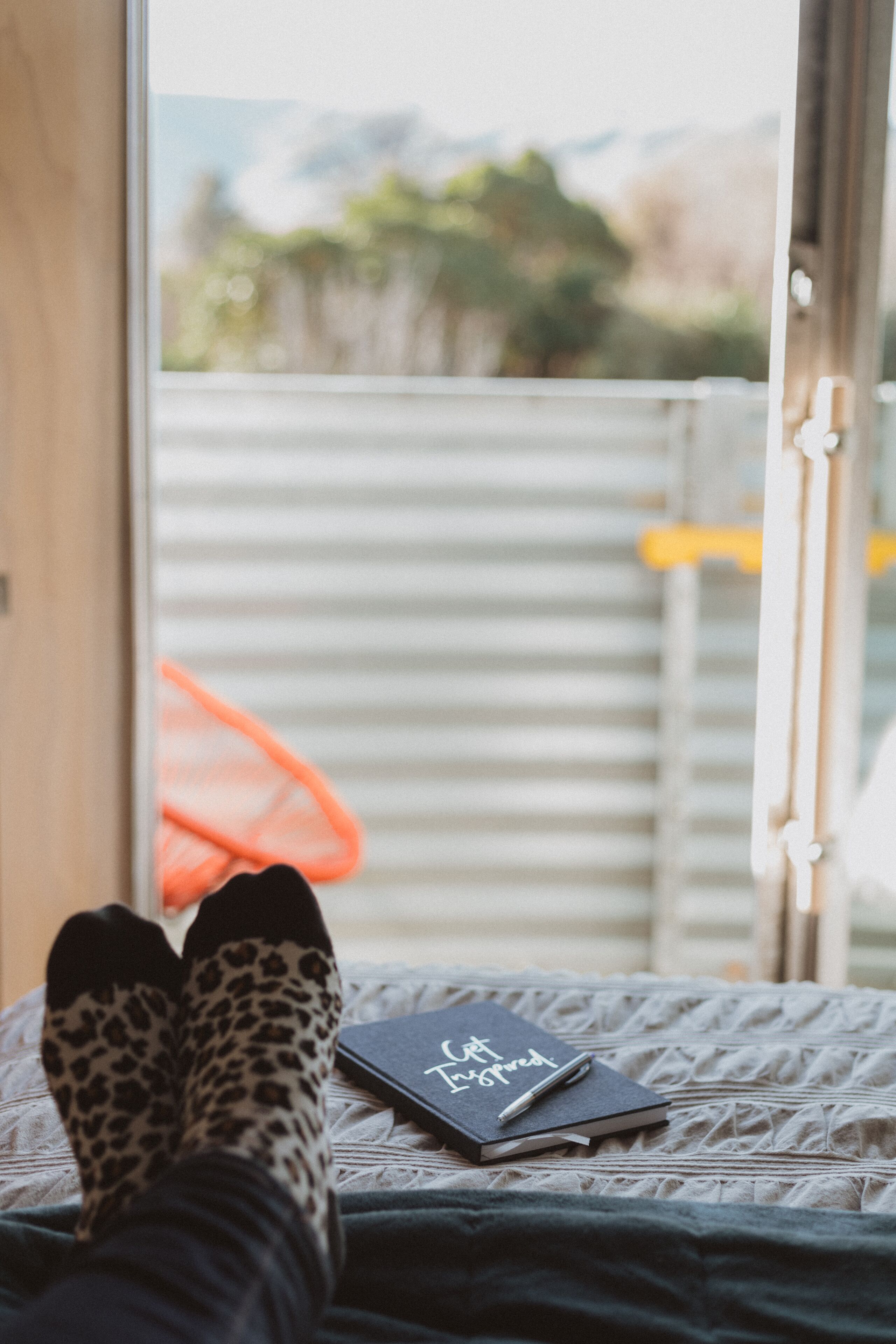 Una cama con un cuaderno y un bolígrafo, una ventana con vistas a árboles y montañas, y un par de calcetines de estampado de leopardo.