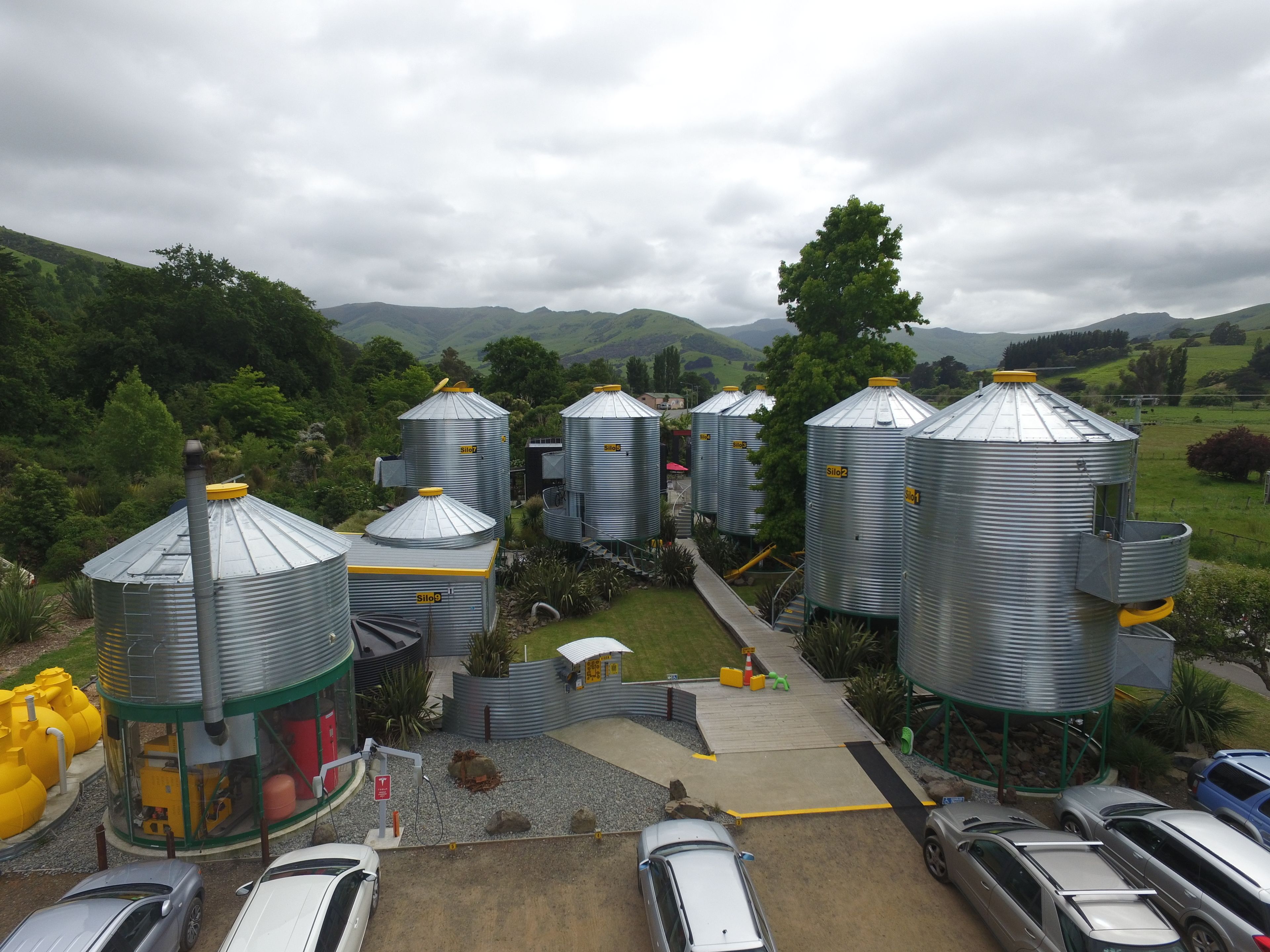 A rural setting with multiple grain silos, a paved area with cars, and a hilly landscape in the background.