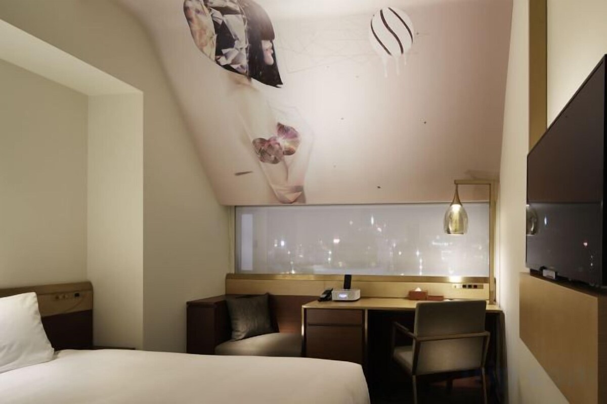 A hotel room with a bed, a desk, and a chair. There is a window with a view of the city.