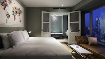 A modern hotel room with a large bed, a seating area with a view of the city, and a bathroom with a bathtub and sink.